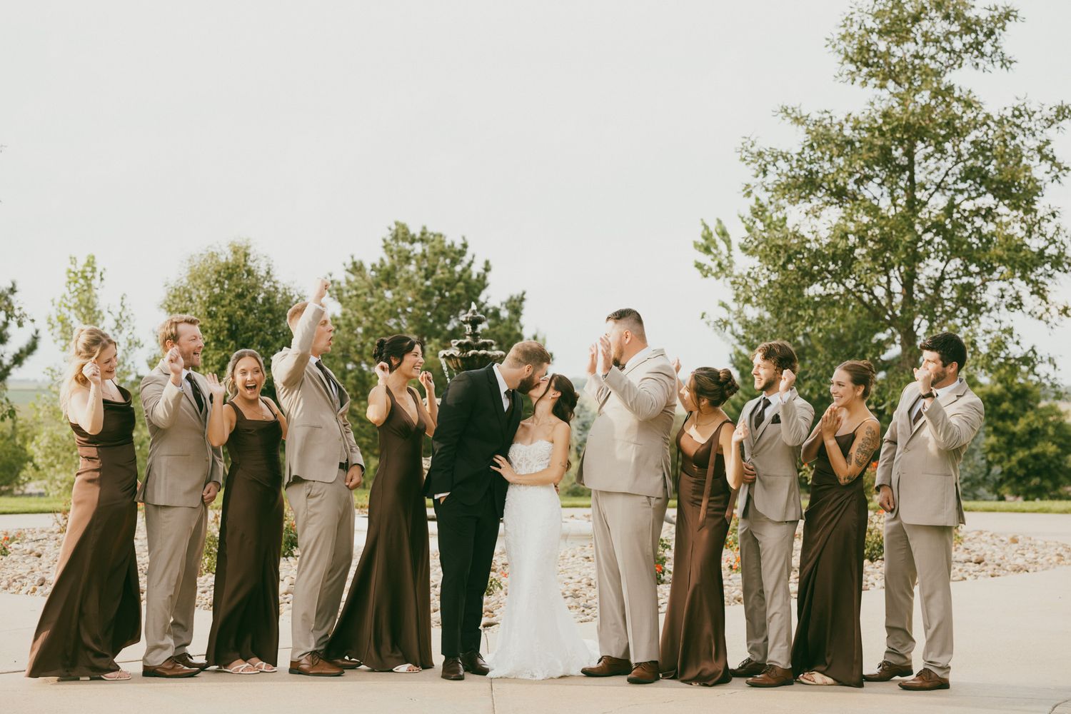 Wedding party celebrating with bride and groom kiss, bridesmaids in dark gowns and groomsmen in tan suits.
