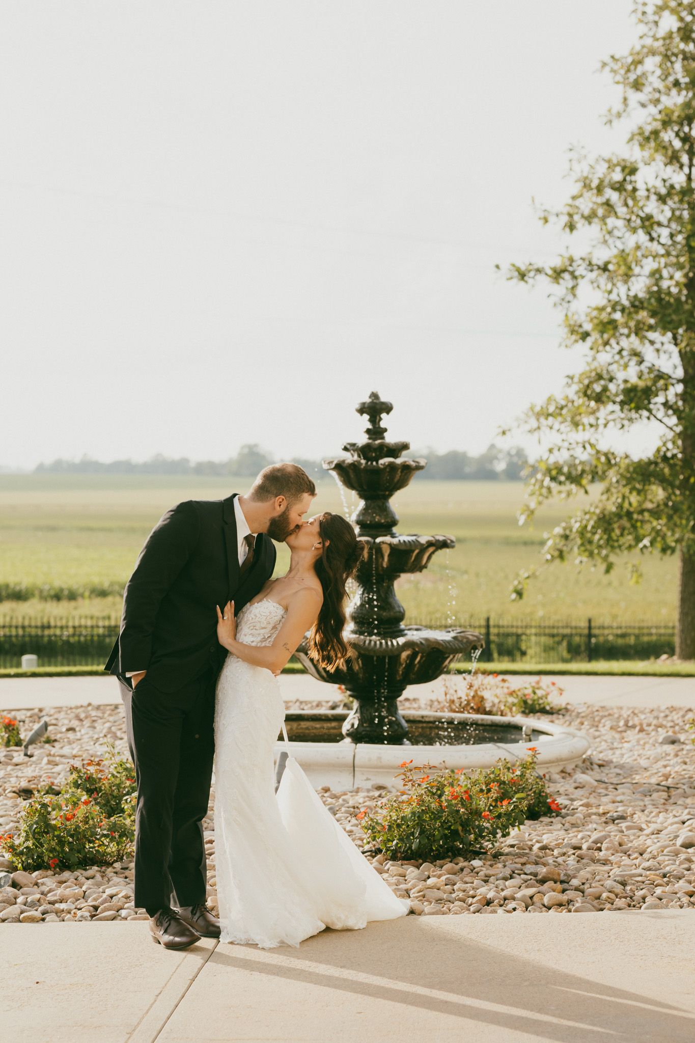 Bride and groom share romantic kiss beside elegant stone fountain at golden hour outdoor venue.