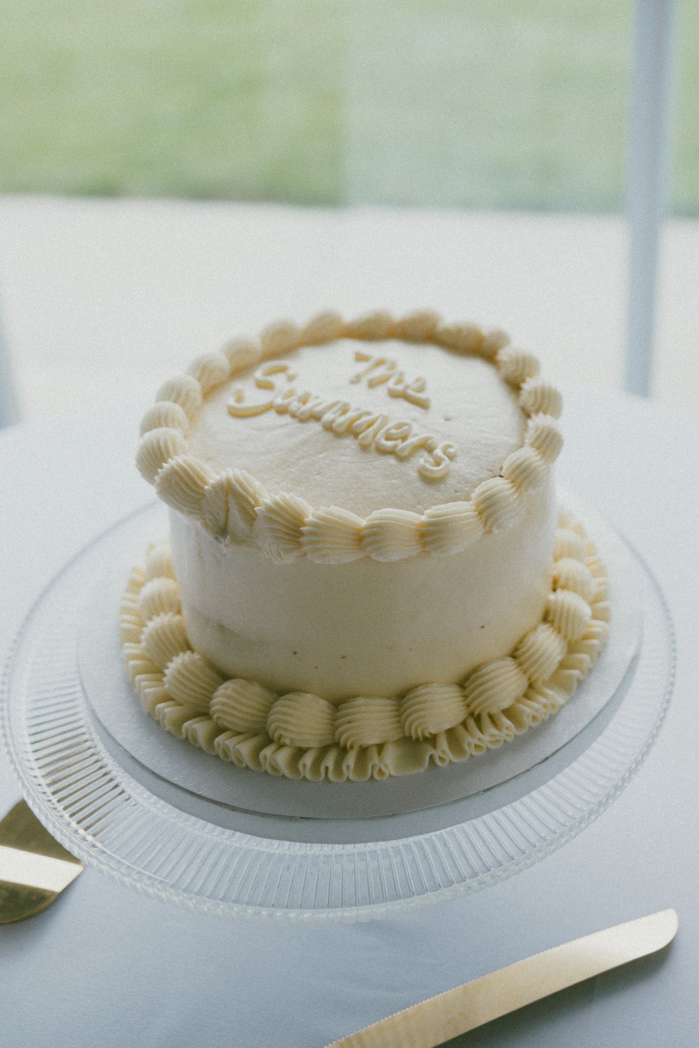 Elegant white wedding cake with cream piping and "The Summers" inscription on a glass cake stand.