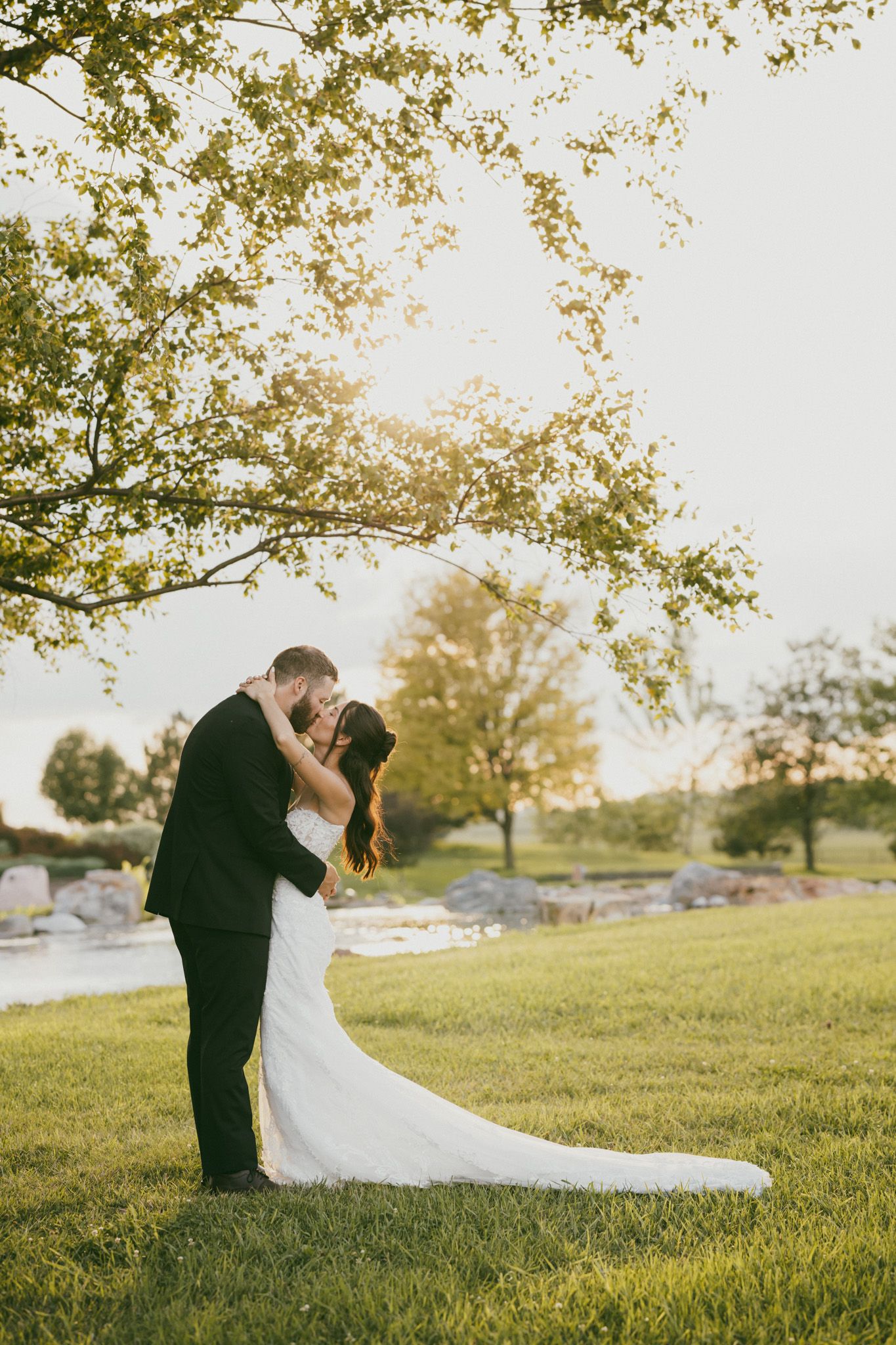 Bride and groom share a romantic kiss under a tree at golden hour during their wedding.