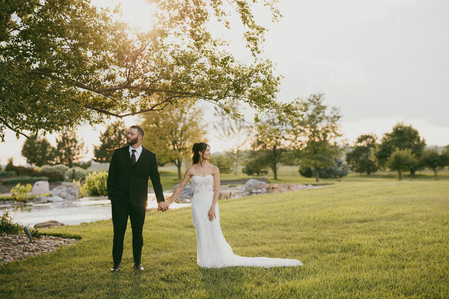 Bride and groom holding hands in sunlit green meadow, bride in white lace gown with train, groom in black tuxedo.
