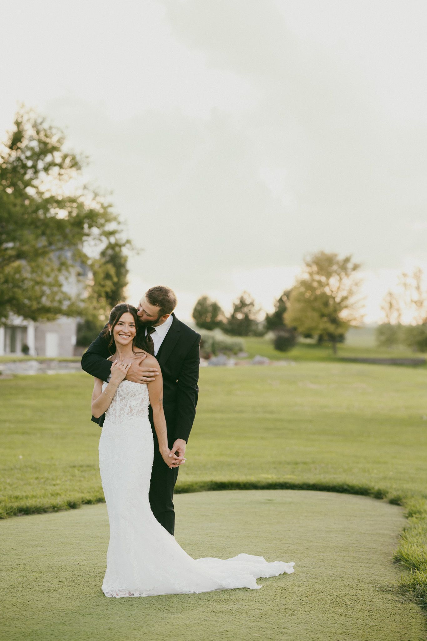 Bride and groom posing on lush green golf course, groom in black tux kissing bride wearing elegant white lace gown.