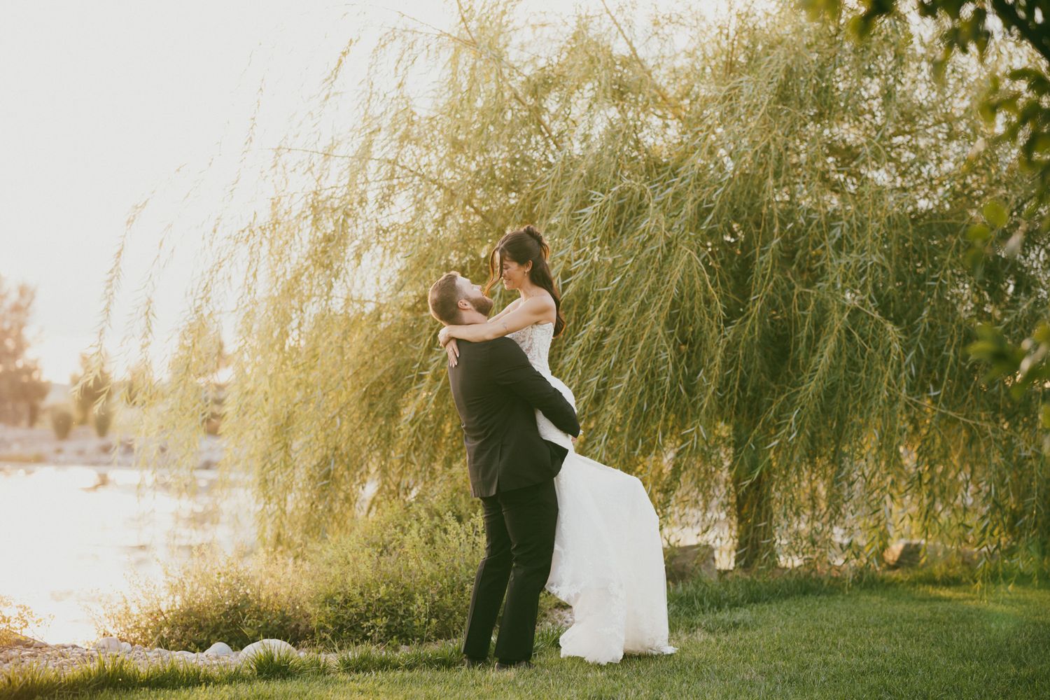 Bride and groom embracing romantically under weeping willow tree by a lake at golden hour.
