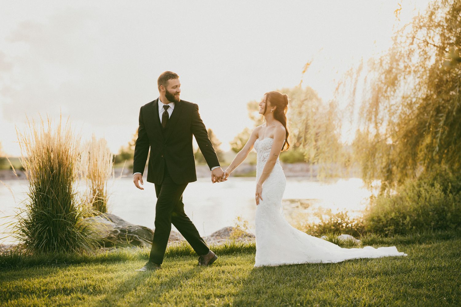 Newlyweds walk hand in hand near a sunlit lake with a weeping willow tree in the background.