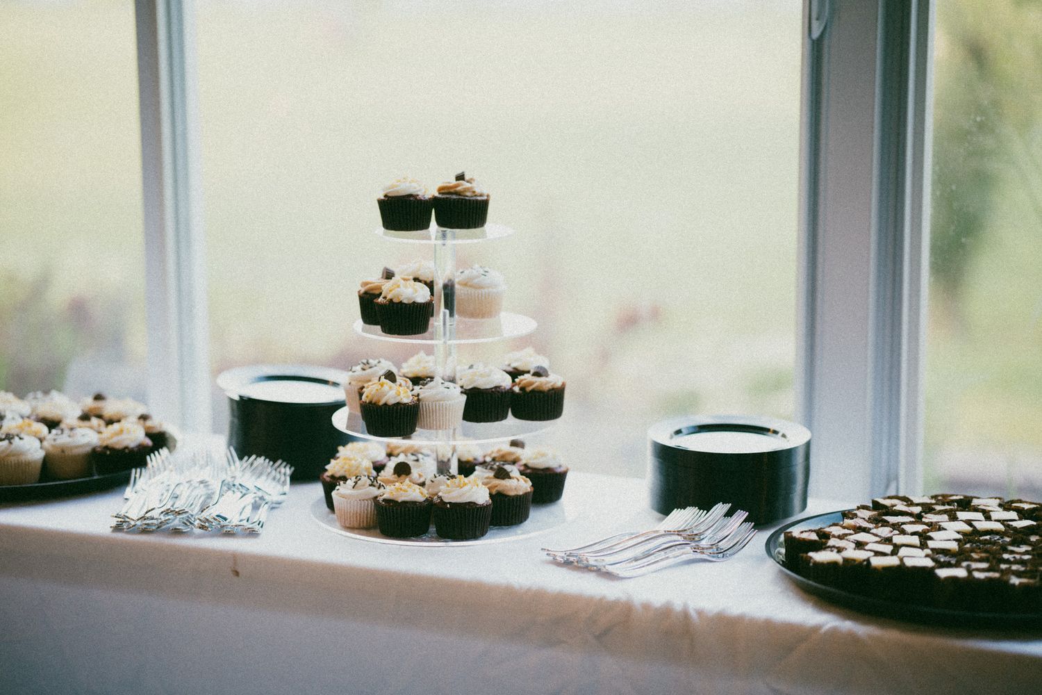 Elegant wedding dessert table with tiered cupcake stand and decorated cookies displayed near window.