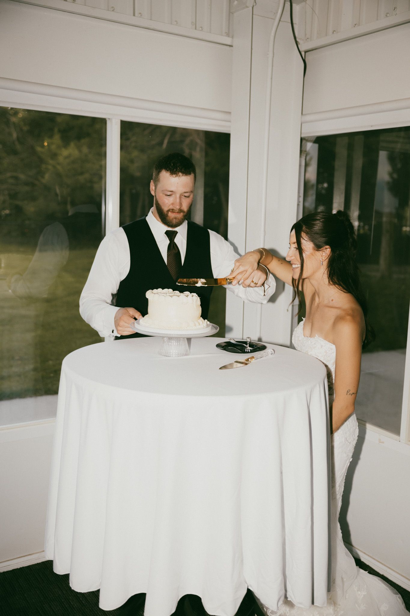 Bride and groom cut their simple white wedding cake together at a white linen-covered table.