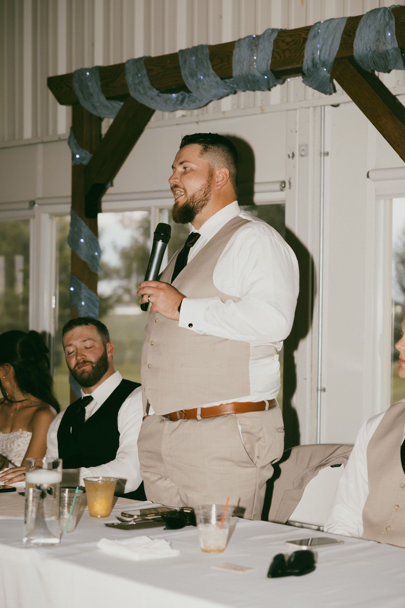 A bearded groomsman in a tan vest delivers a wedding speech at a reception while holding a microphone.