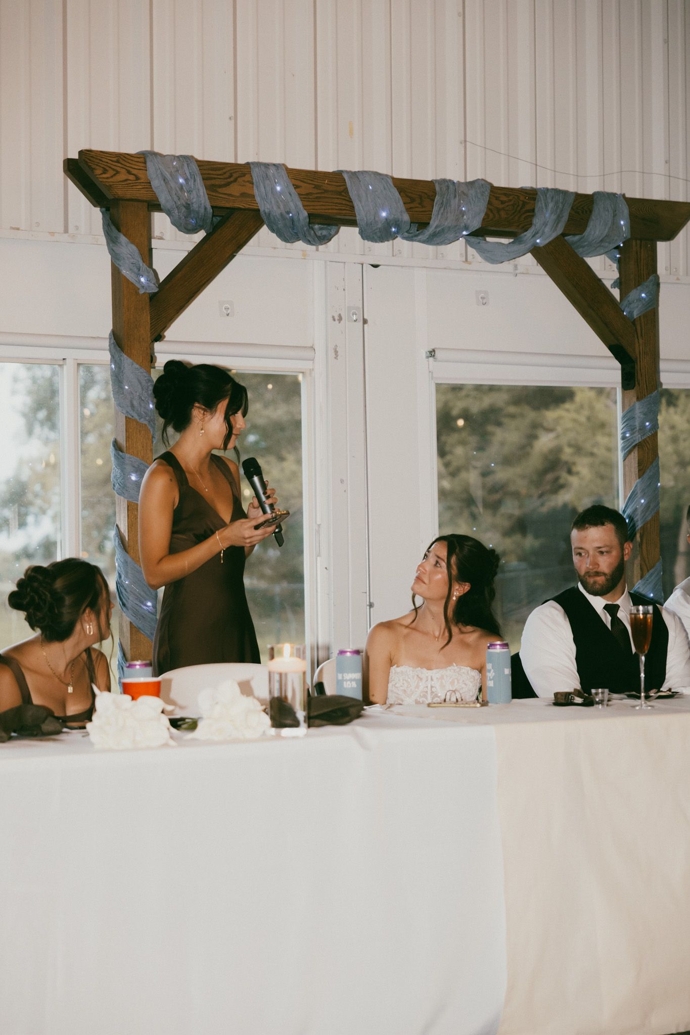 Bridesmaid gives a heartfelt speech at the wedding reception as the bride smiles joyfully.