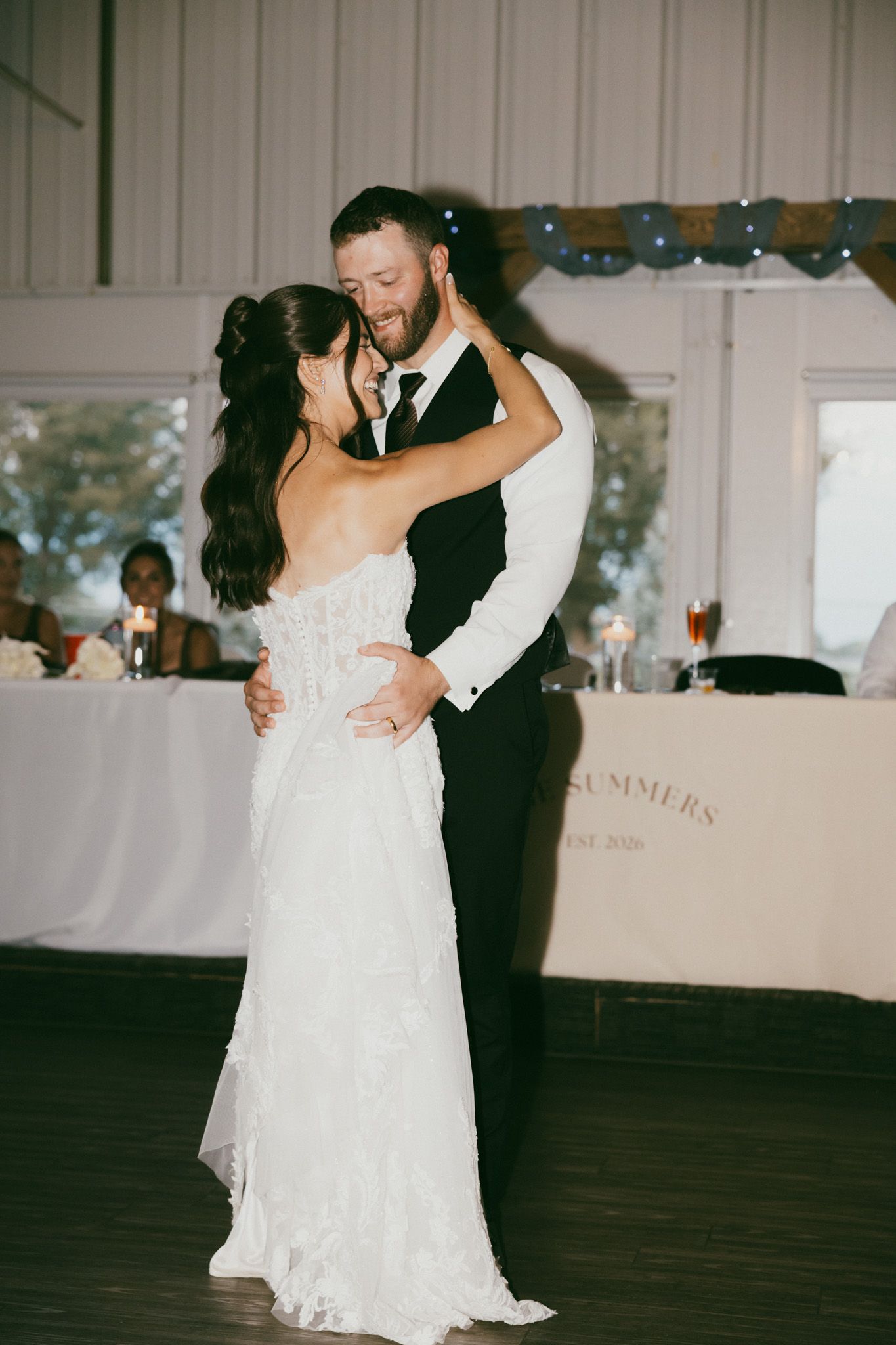 Bride and groom share their first dance at wedding reception, smiling intimately in elegant white gown and dark suit.