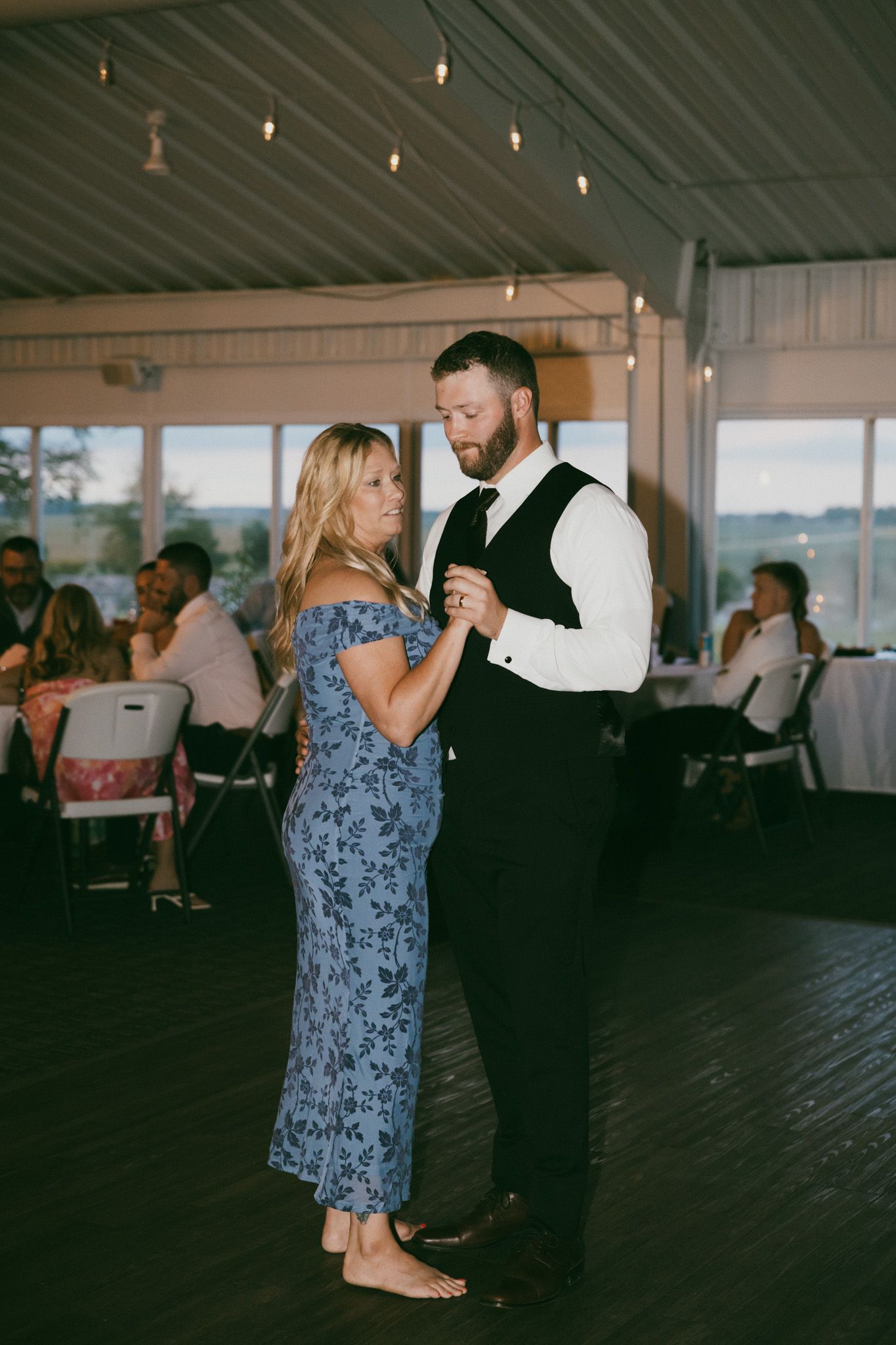 A groom in a black vest dances with a woman in a blue floral dress at a wedding reception with string lights.