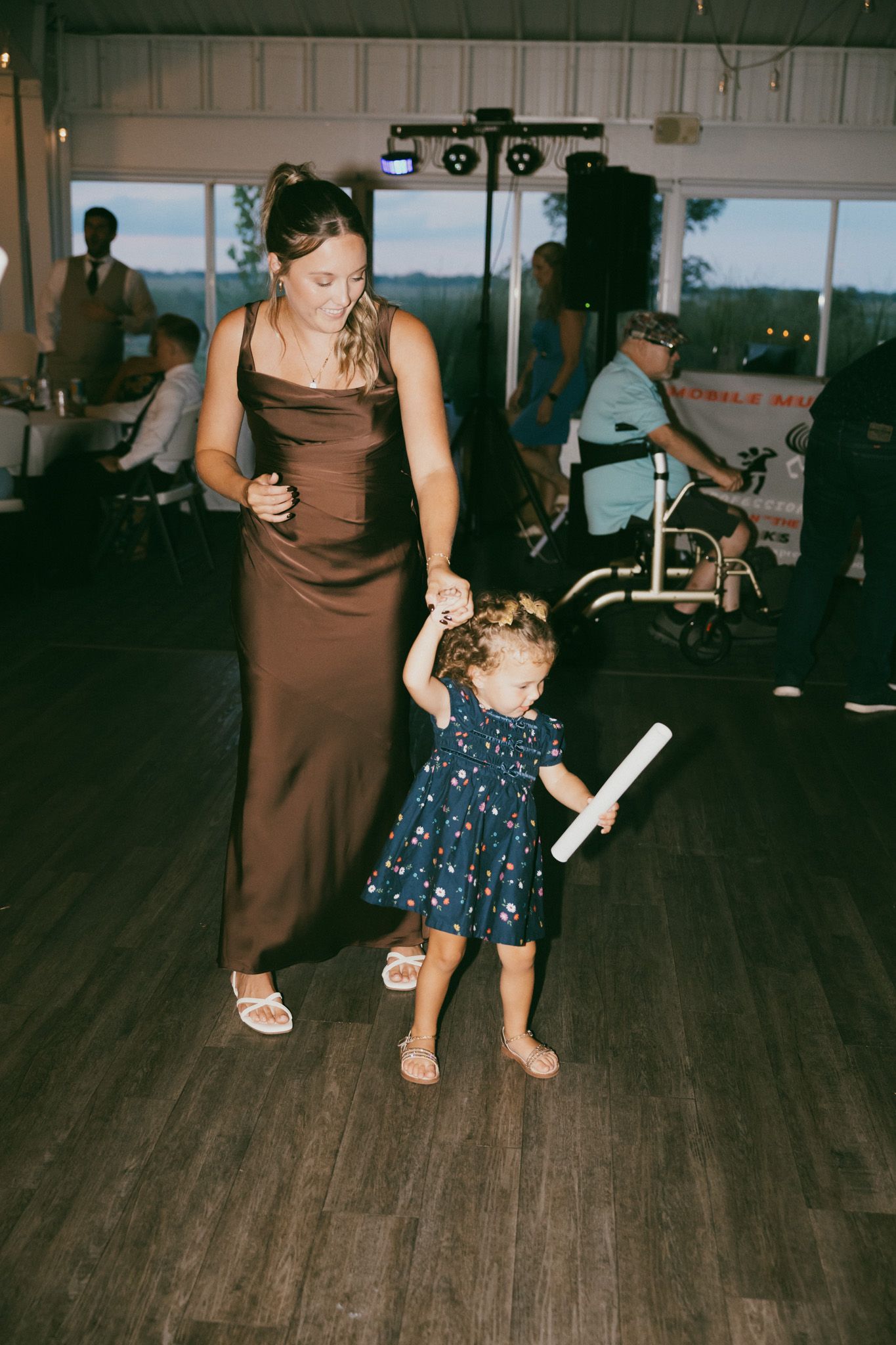 A woman in a brown dress dances with a toddler girl in a floral dress at a wedding reception.