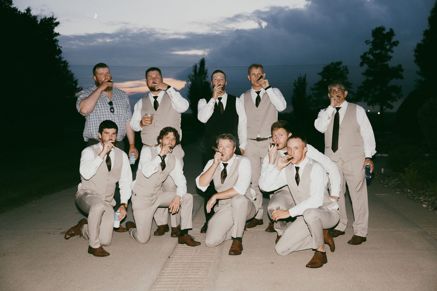Groomsmen in white shirts and tan suits pose playfully outdoors at dusk during a wedding celebration.