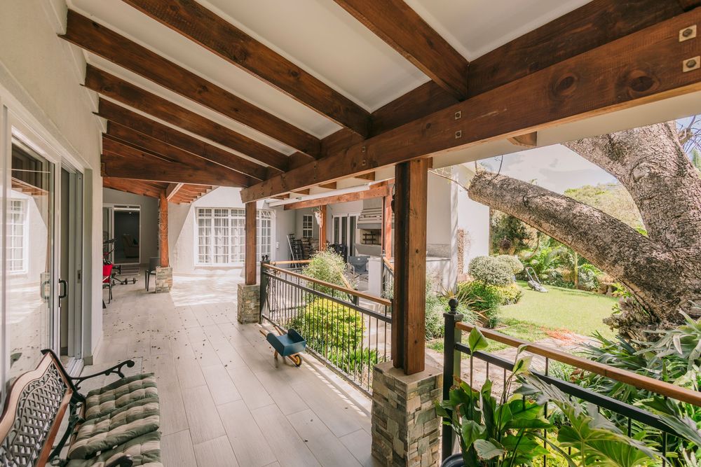 Elegant covered porch with rustic wooden beam ceiling, white columns, and lush garden views beyond decorative iron railing.
