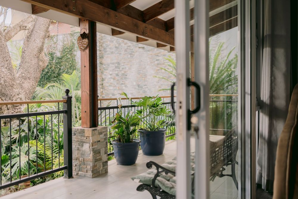 Covered balcony with blue potted plants, stone pillars, black iron railing, and lush tropical garden views.
