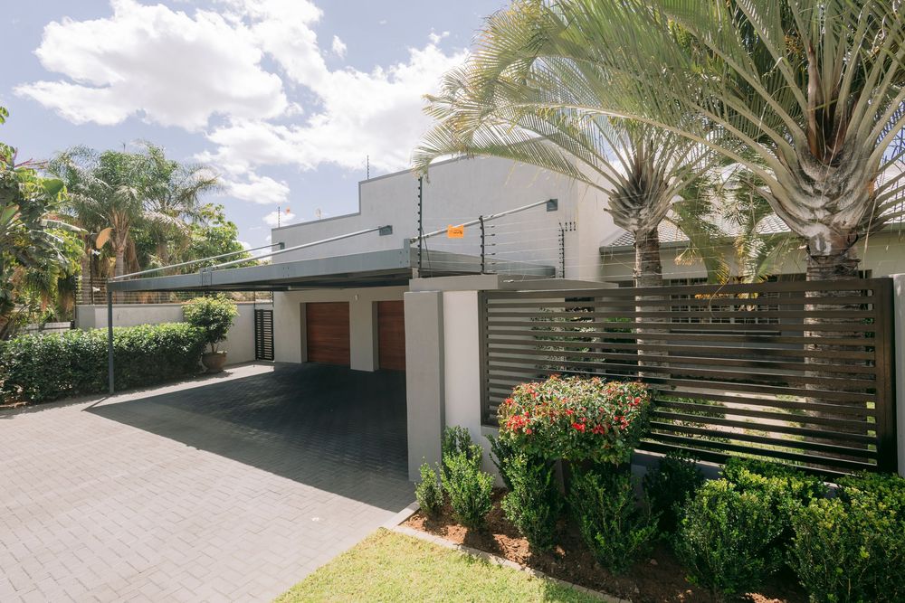 Modern white home with carport, timber screen fence, tropical palms, and manicured gardens on a paved driveway.