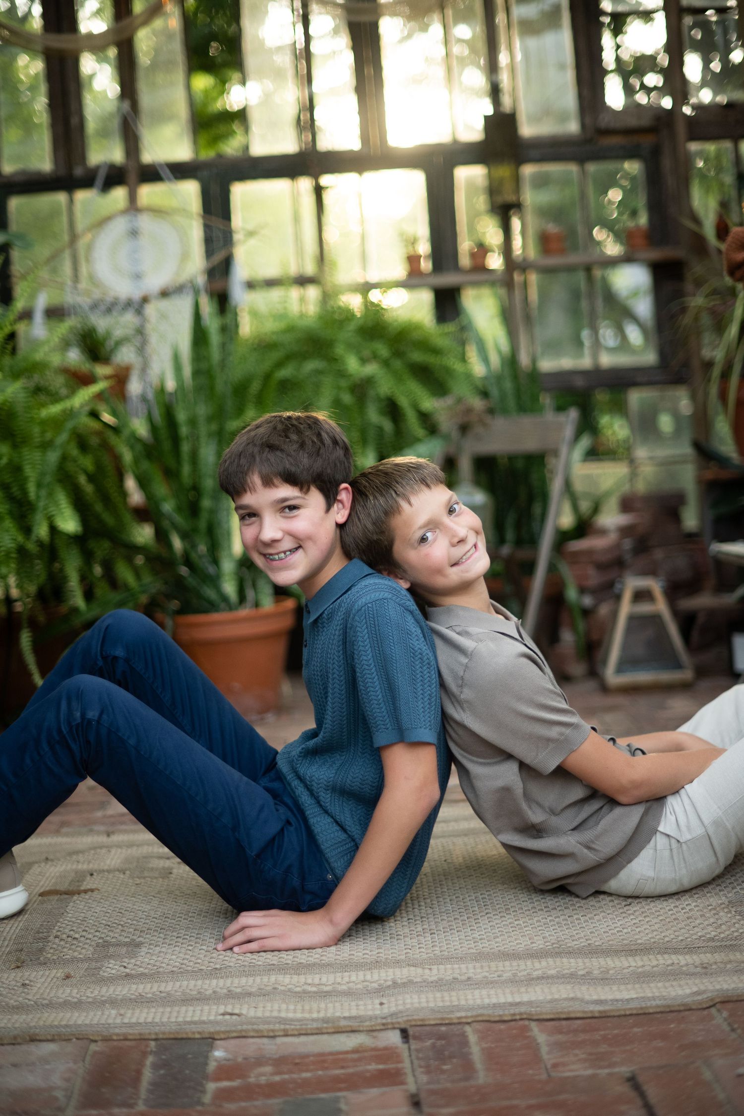 Two smiling boys sitting back-to-back on a rug in a sunlit greenhouse surrounded by lush green plants.