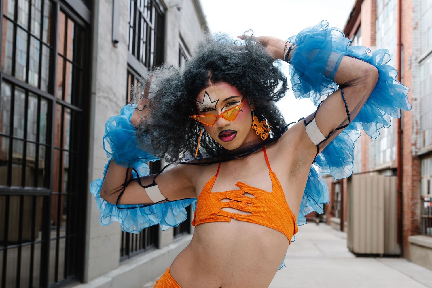 Drag performer in orange bikini top with blue feather accessories and curly wig posing in urban alleyway.