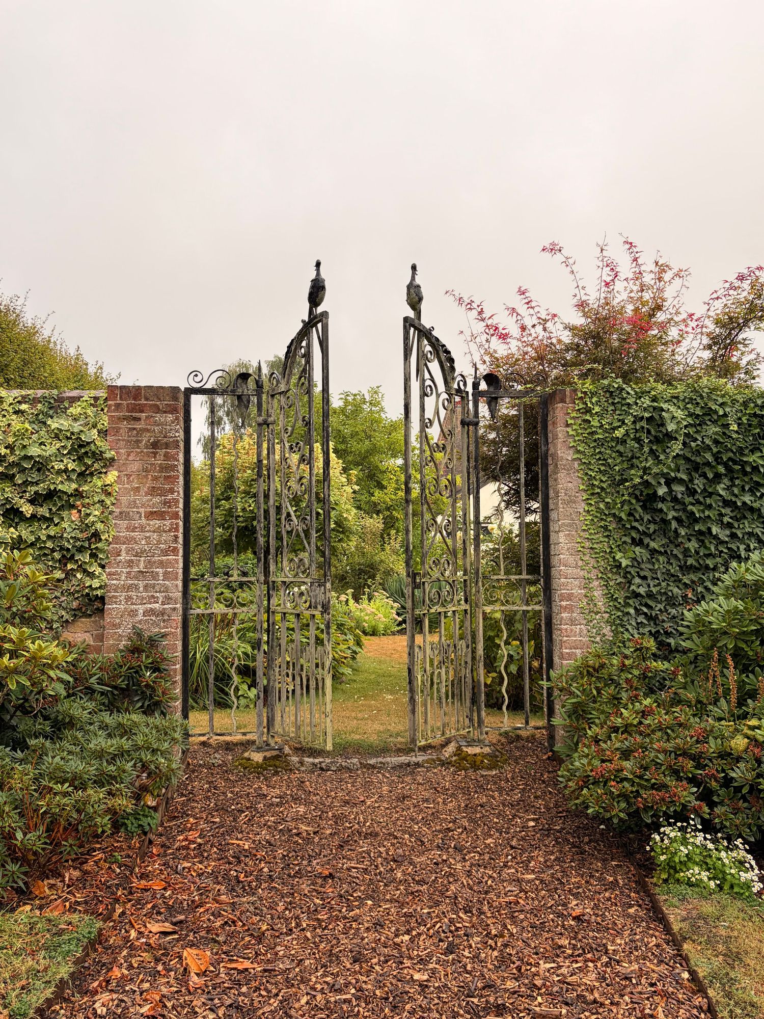 Ornate wrought iron garden gate with bird finials set in brick wall, opening to lush green garden with autumn leaves on path.