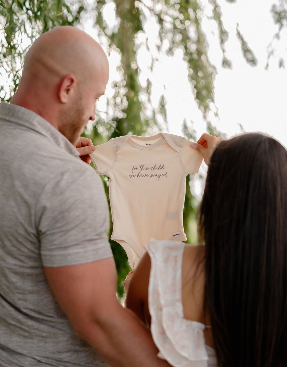 Couple holding up a white baby onesie reading 'for this child we have prayed' outdoors.