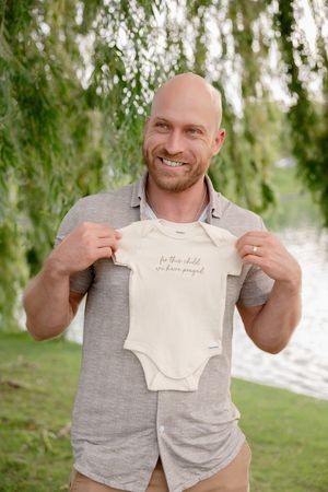 Smiling bald man holding a baby onesie outdoors near a willow tree and lake, announcing pregnancy.