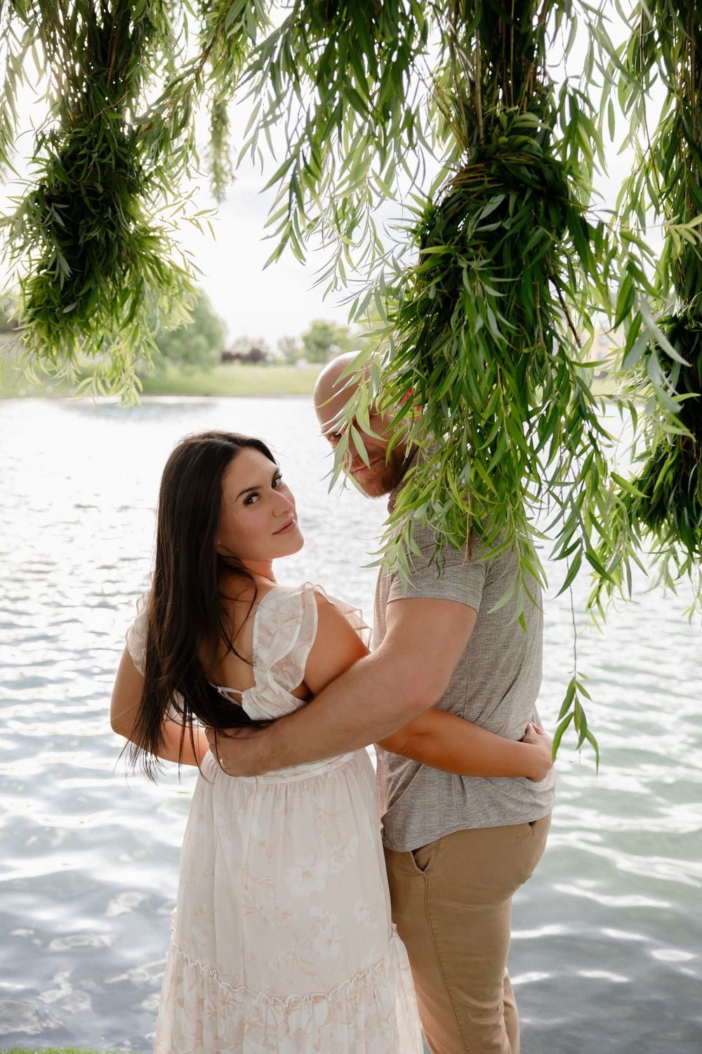 Romantic couple embracing under weeping willow tree branches by a serene lake, woman in white dress looking back.