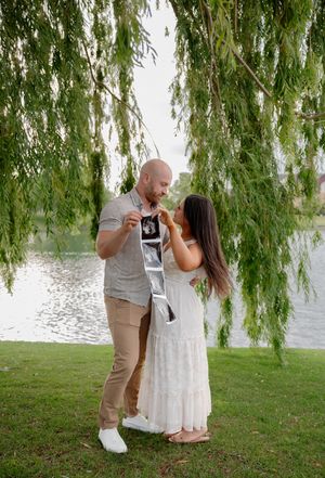 Couple holding ultrasound photos under a weeping willow tree by a lake, celebrating their pregnancy announcement outdoors.