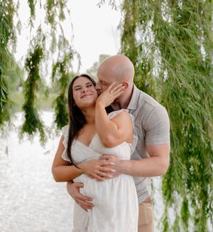 Couple sharing a tender moment under weeping willow branches by a lake, pregnant woman in white dress smiling.