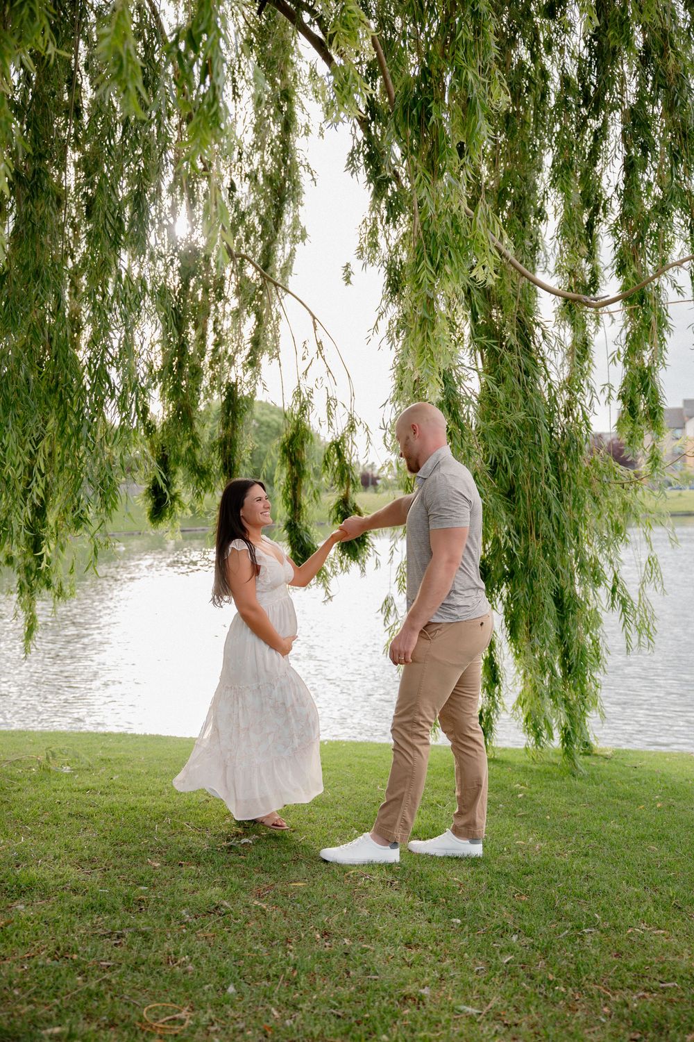 Pregnant woman in white dress and bald man hold hands by a willow tree near a lake in a romantic outdoor setting.