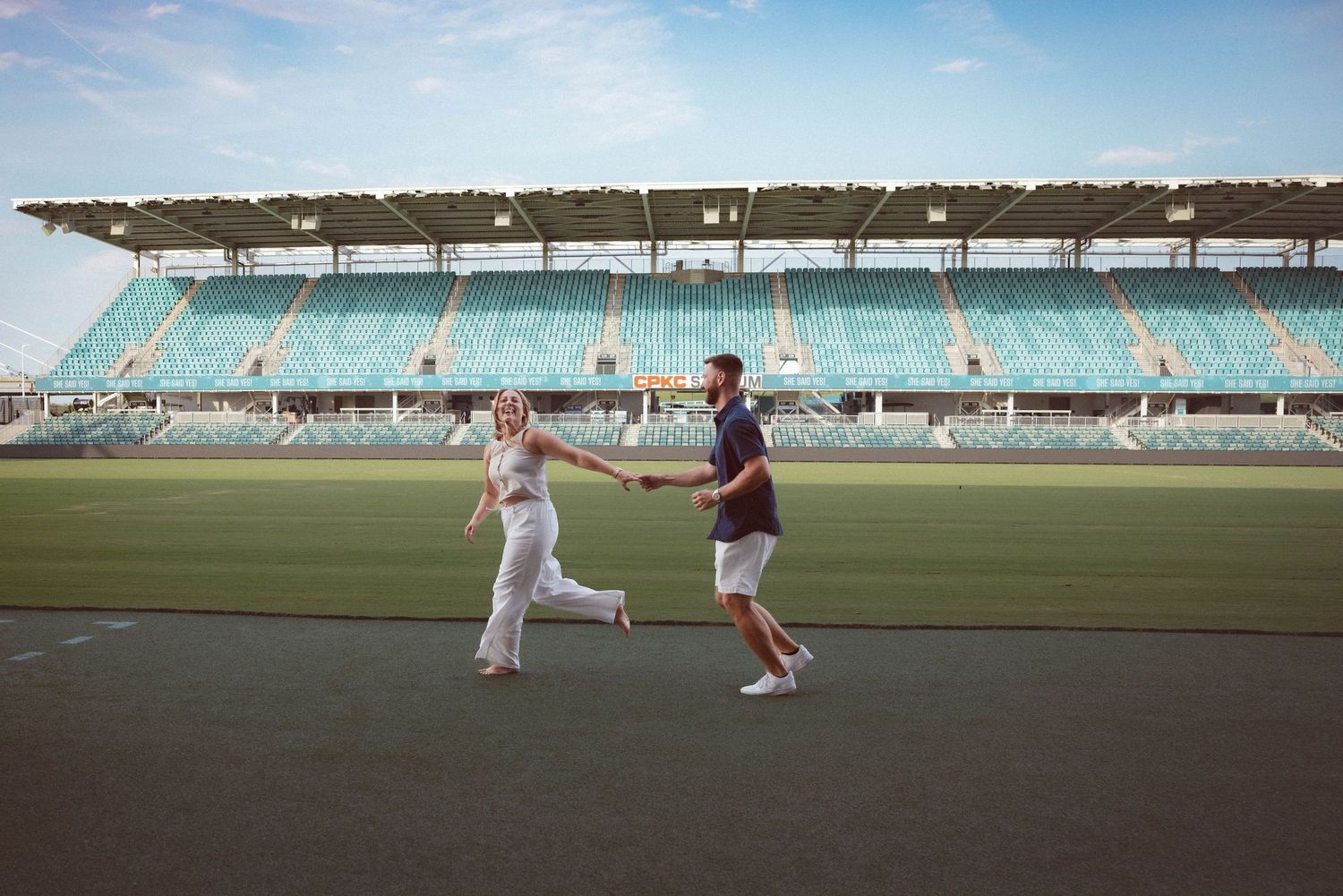 A newly engaged man and woman run across the KC Current soccer field following their surprise proposal at CPKC stadium.