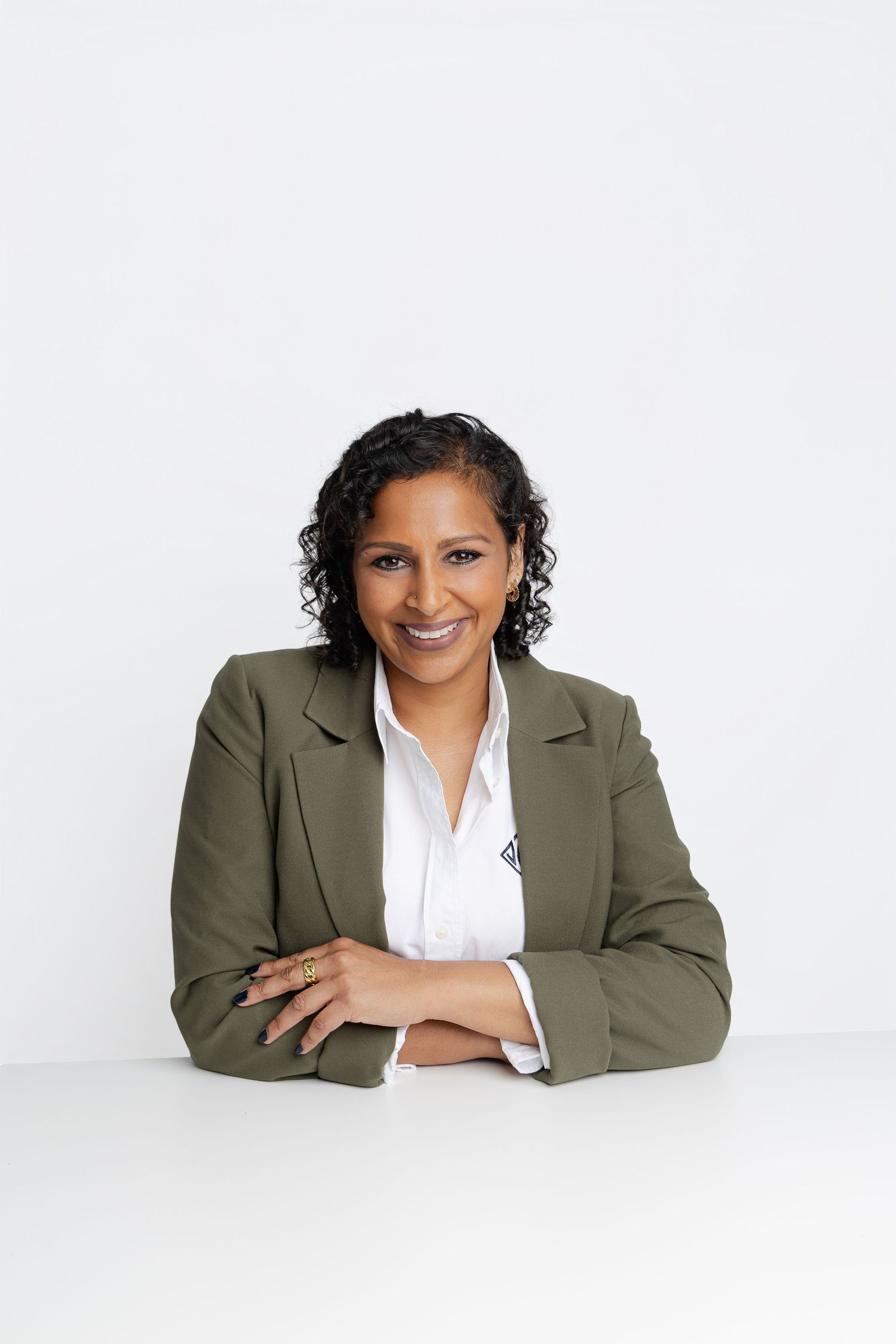 Professional woman in olive blazer smiling with arms crossed at white desk against light background.