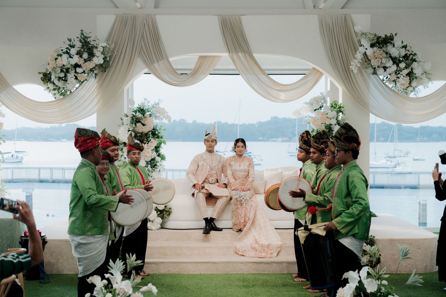 Traditional Malay wedding ceremony with couple seated on stage, performers in green, floral decor, and waterfront backdrop.