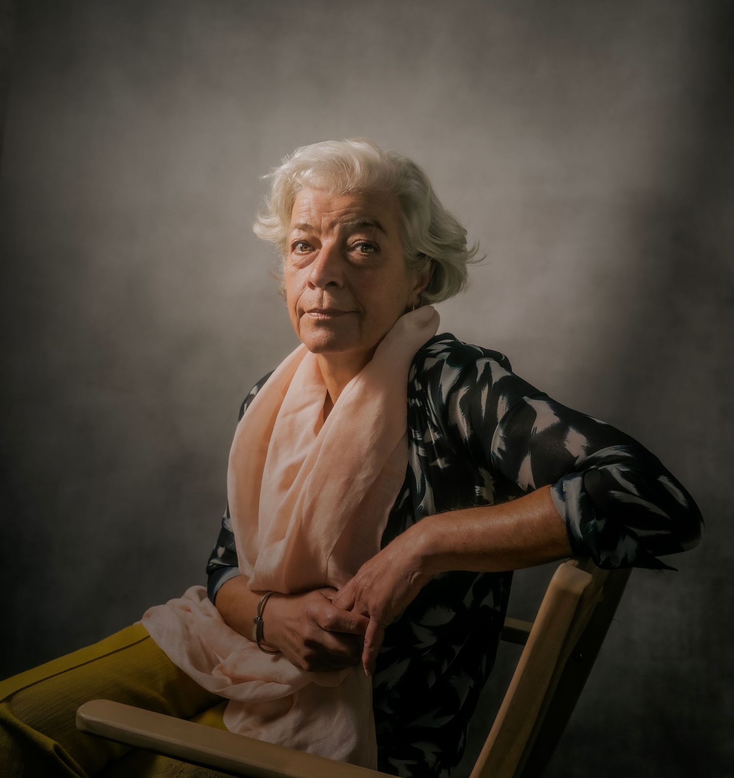 Elderly woman with white hair wearing a floral blouse and pink scarf, seated on a wooden chair against a gray backdrop.
