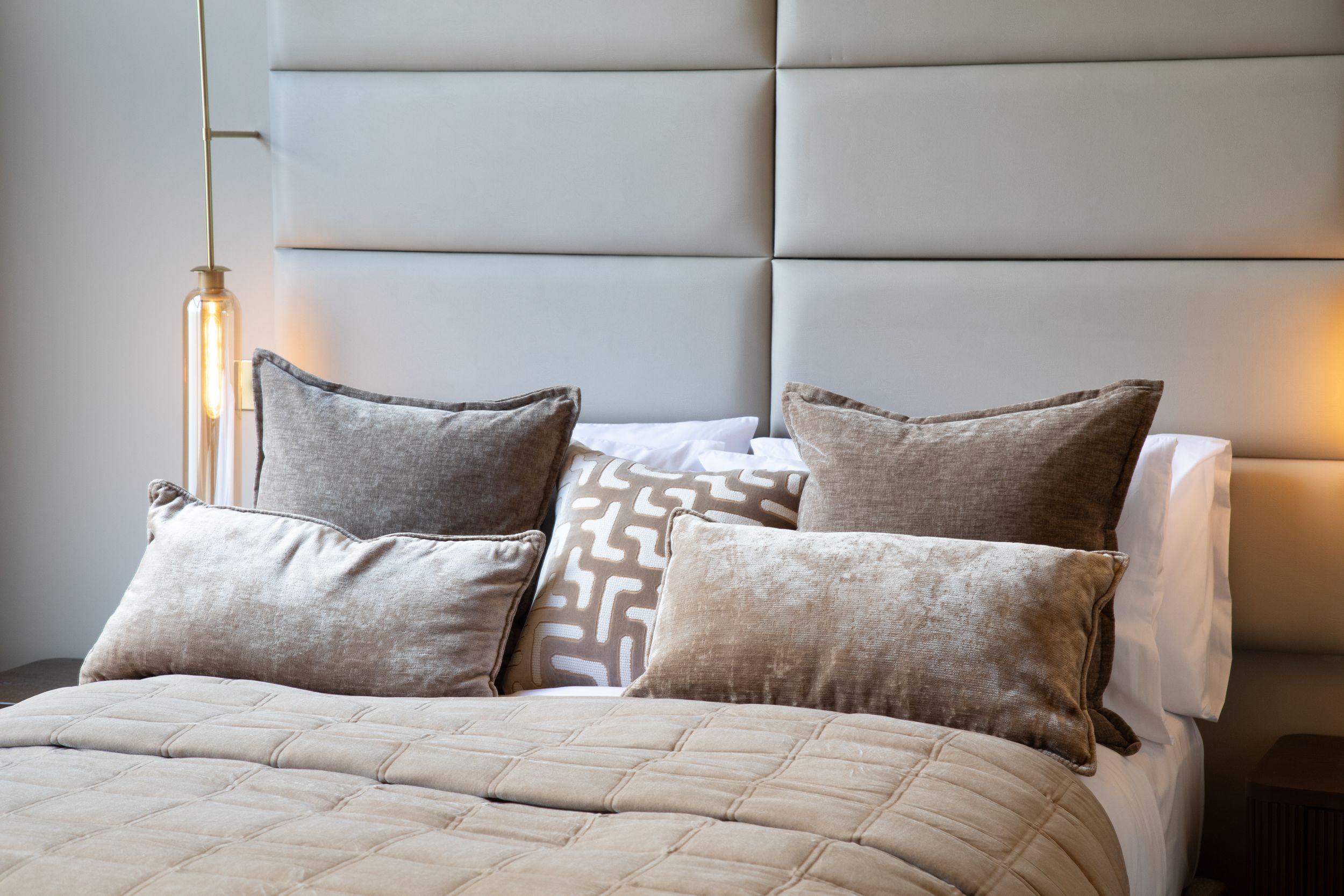 Elegant bed with quilted cream bedding, taupe velvet pillows, and a tall upholstered headboard with warm pendant lighting.