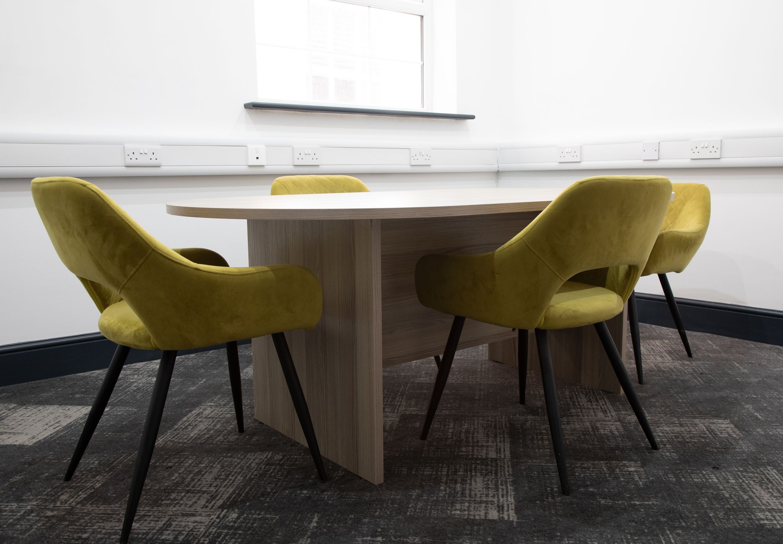 Small modern meeting room with square wooden table surrounded by four olive green velvet chairs.