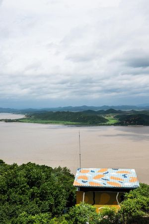Aerial view of a yellow building with colorful roof overlooking a wide muddy river and green hills under cloudy skies.