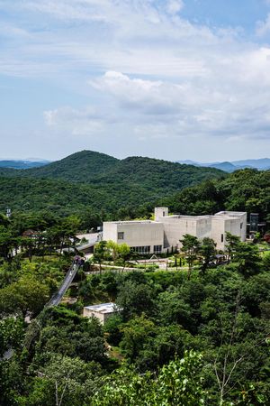 Modern museum building nestled among lush green forested hills under a partly cloudy blue sky in South Korea.