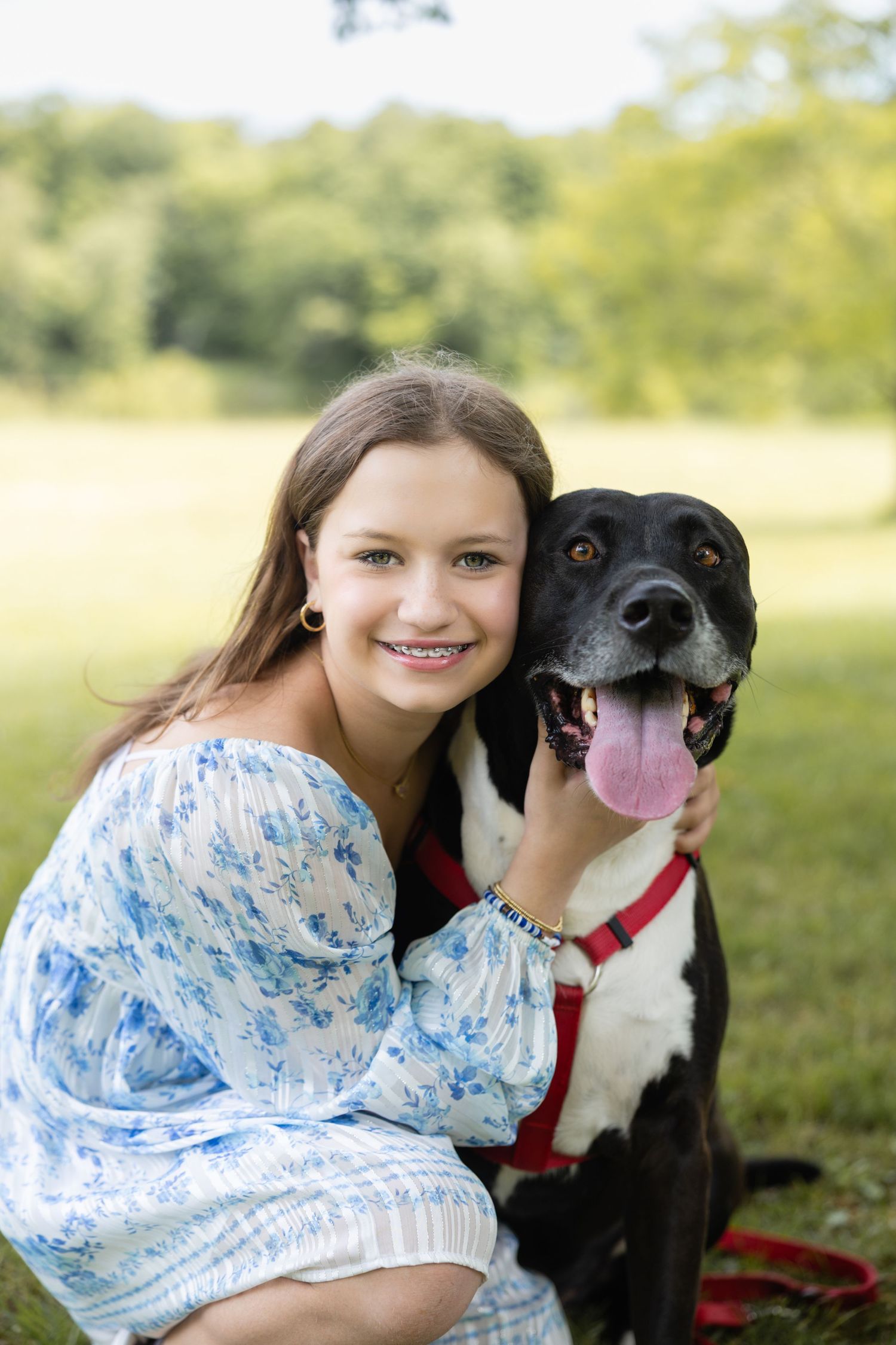 Smiling teenage girl with braces in floral dress hugging a black and white dog with red leash in a green park.
