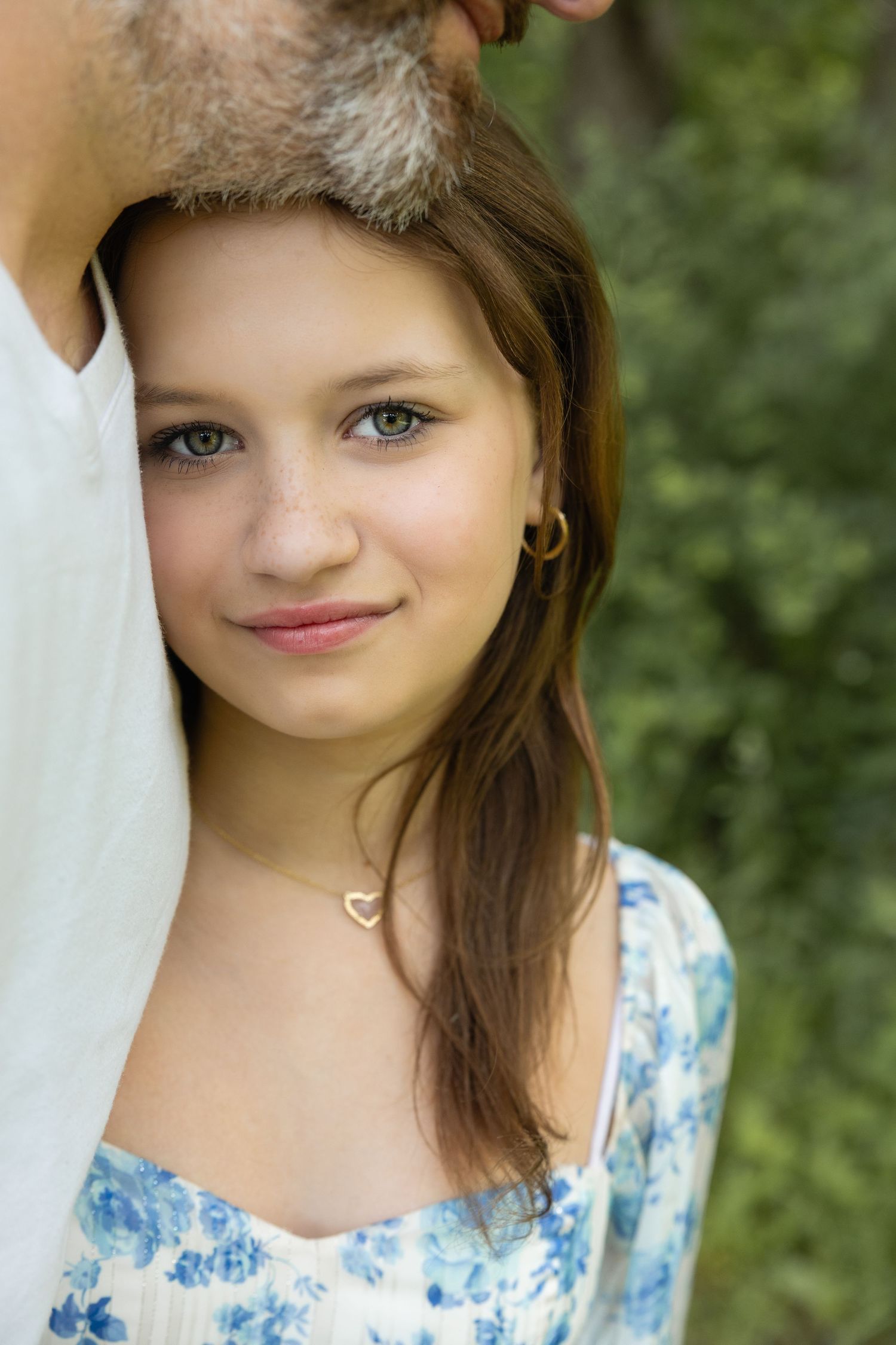 Young woman with blue eyes wearing a floral dress and heart necklace, leaning against a man outdoors.