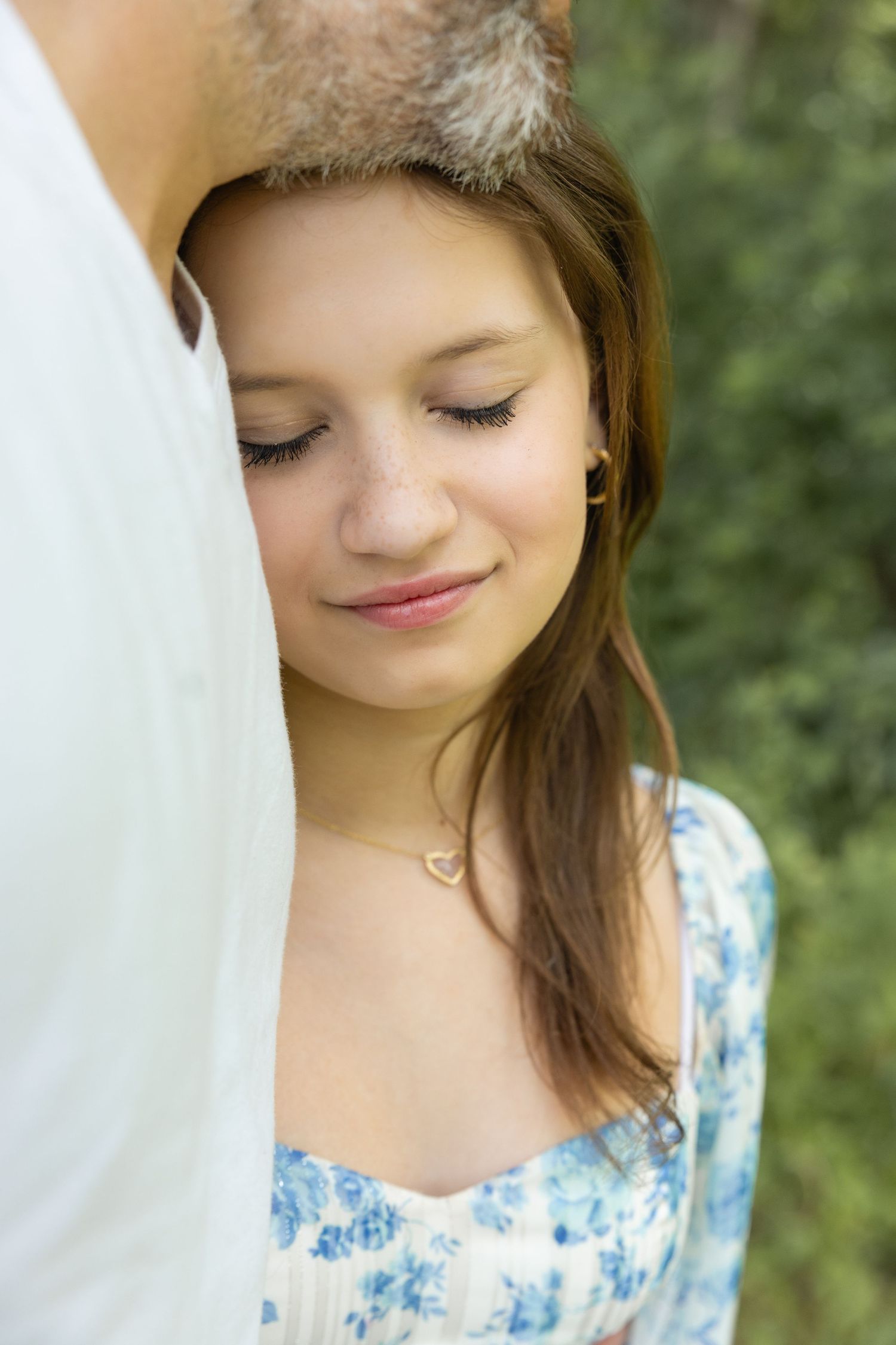 Young woman in floral dress resting her head against a man's shoulder outdoors, smiling softly with eyes downcast.