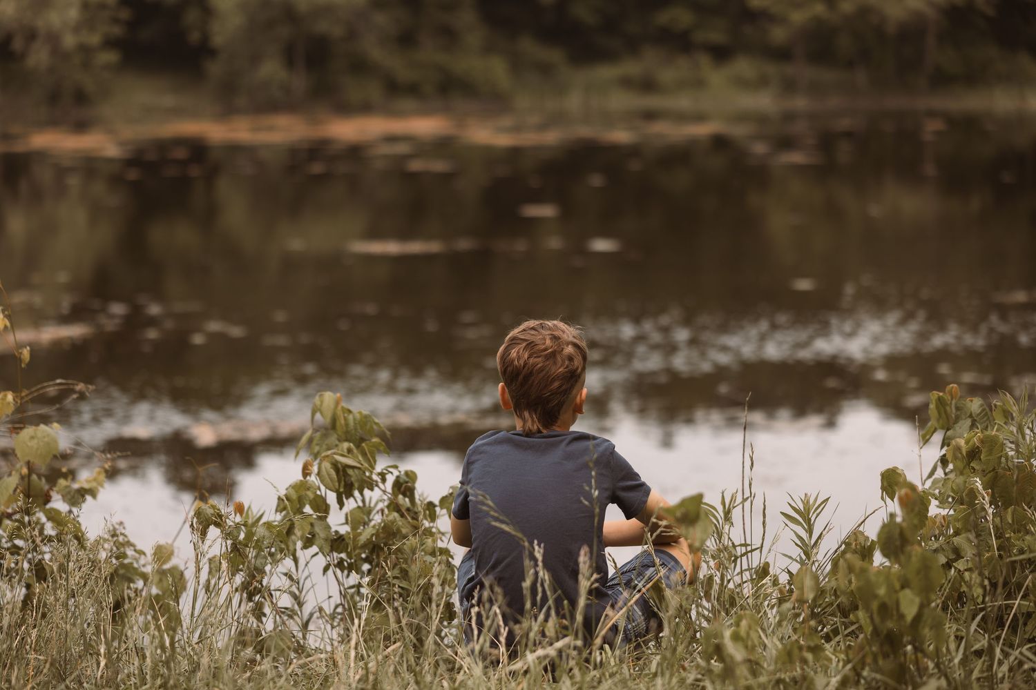 A young boy sits alone in tall grass, gazing peacefully at a calm reflective pond surrounded by lush green trees.