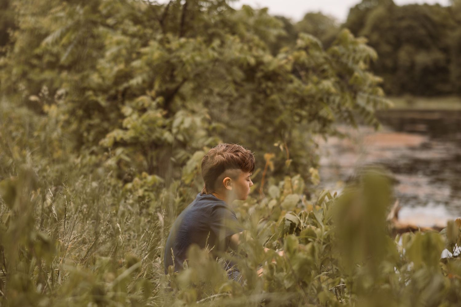 A young man sits among lush green bushes near a tranquil pond surrounded by trees.