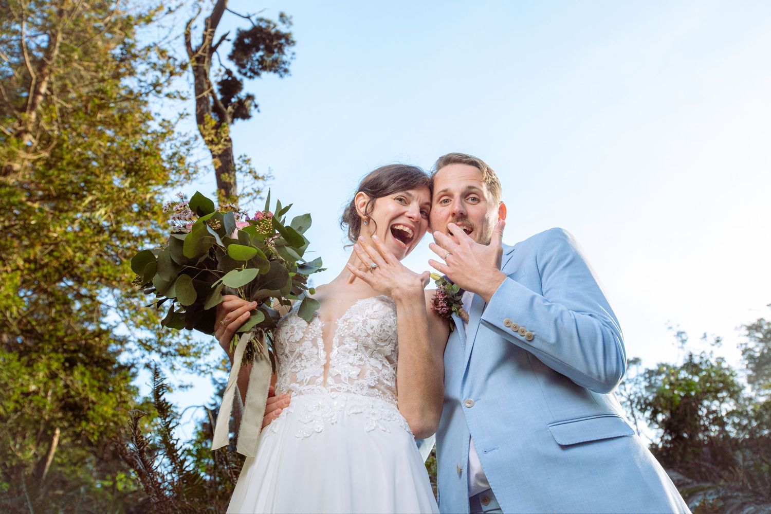 Joyful bride and groom laughing together outdoors, bride holding bouquet, groom in light blue suit on their wedding day.