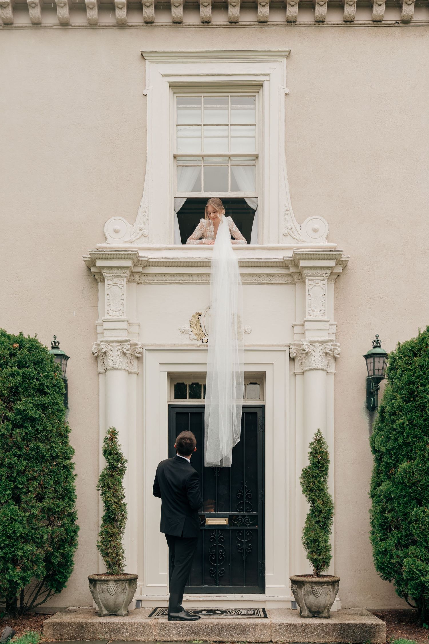 Bride in white gown and veil leans from window above groom in black tux at elegant estate entrance.