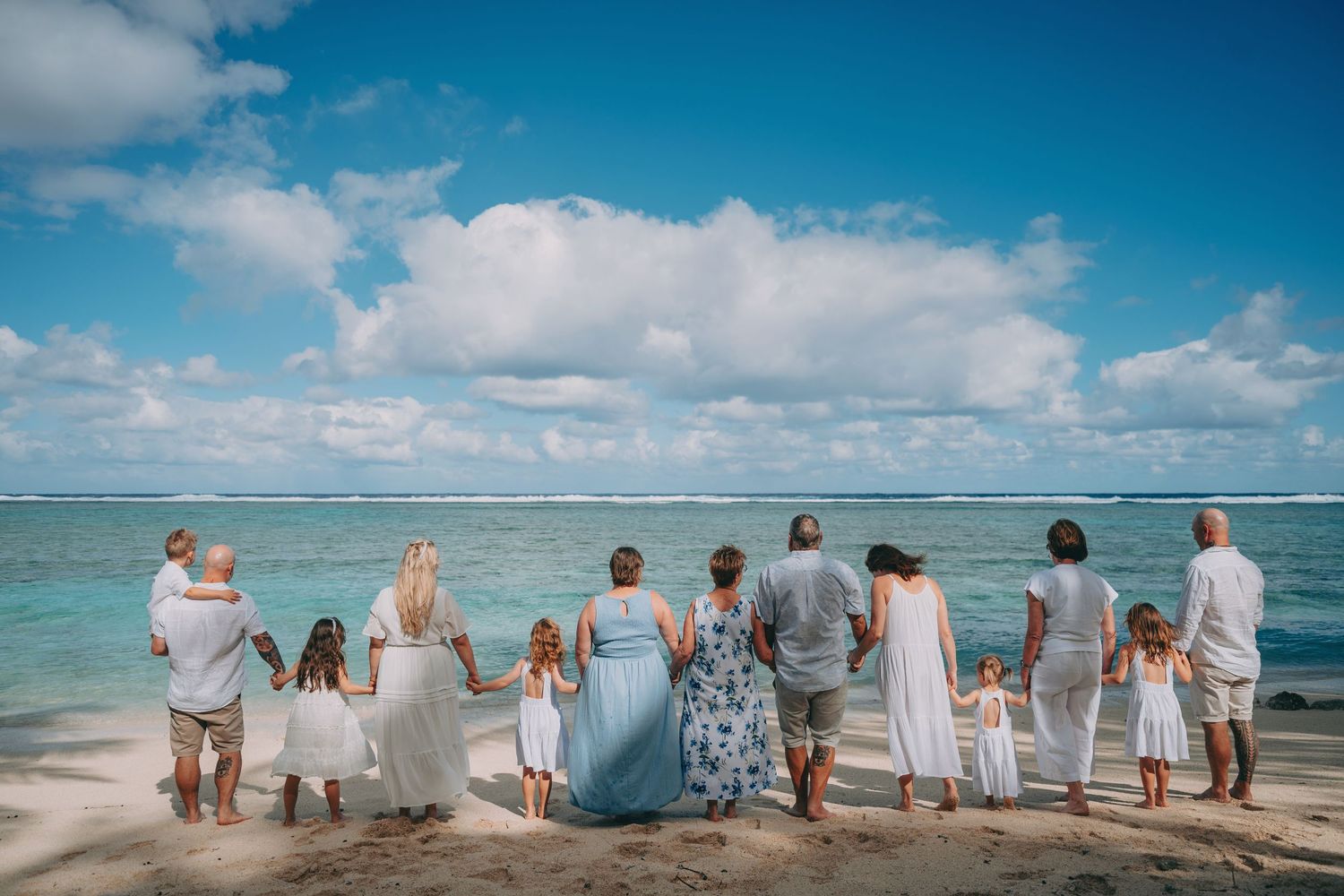 Large family holding hands on a tropical beach, facing turquoise ocean under a bright blue sky with white clouds.