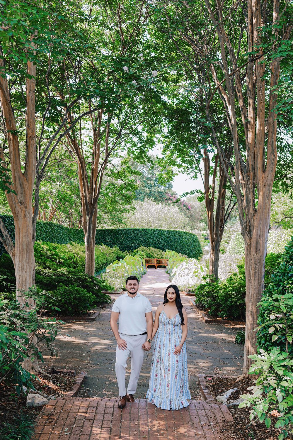 Couple holding hands on a brick garden path lined with tall trees and lush greenery on a sunny day.