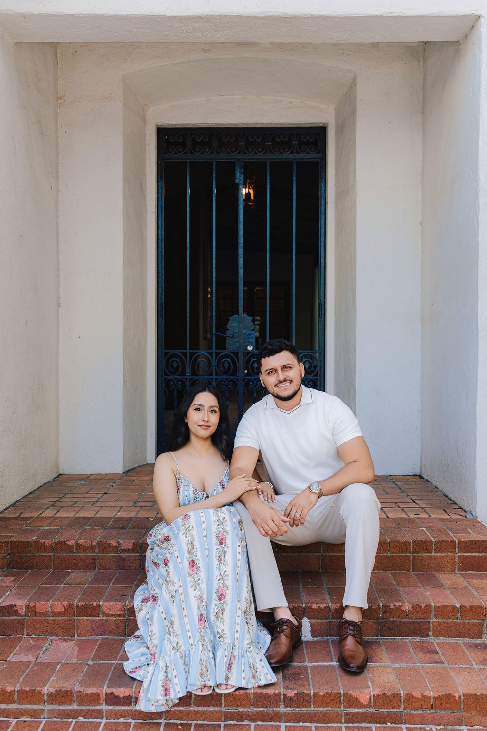 Couple sitting on brick steps in front of ornate iron gate, woman in floral dress, man in white shirt and khakis.