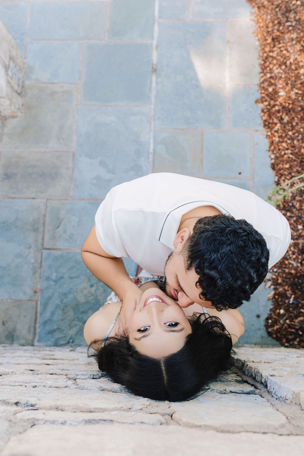 A young couple shares a tender kiss outdoors, with the woman lying on stone pavers and the man leaning over her.