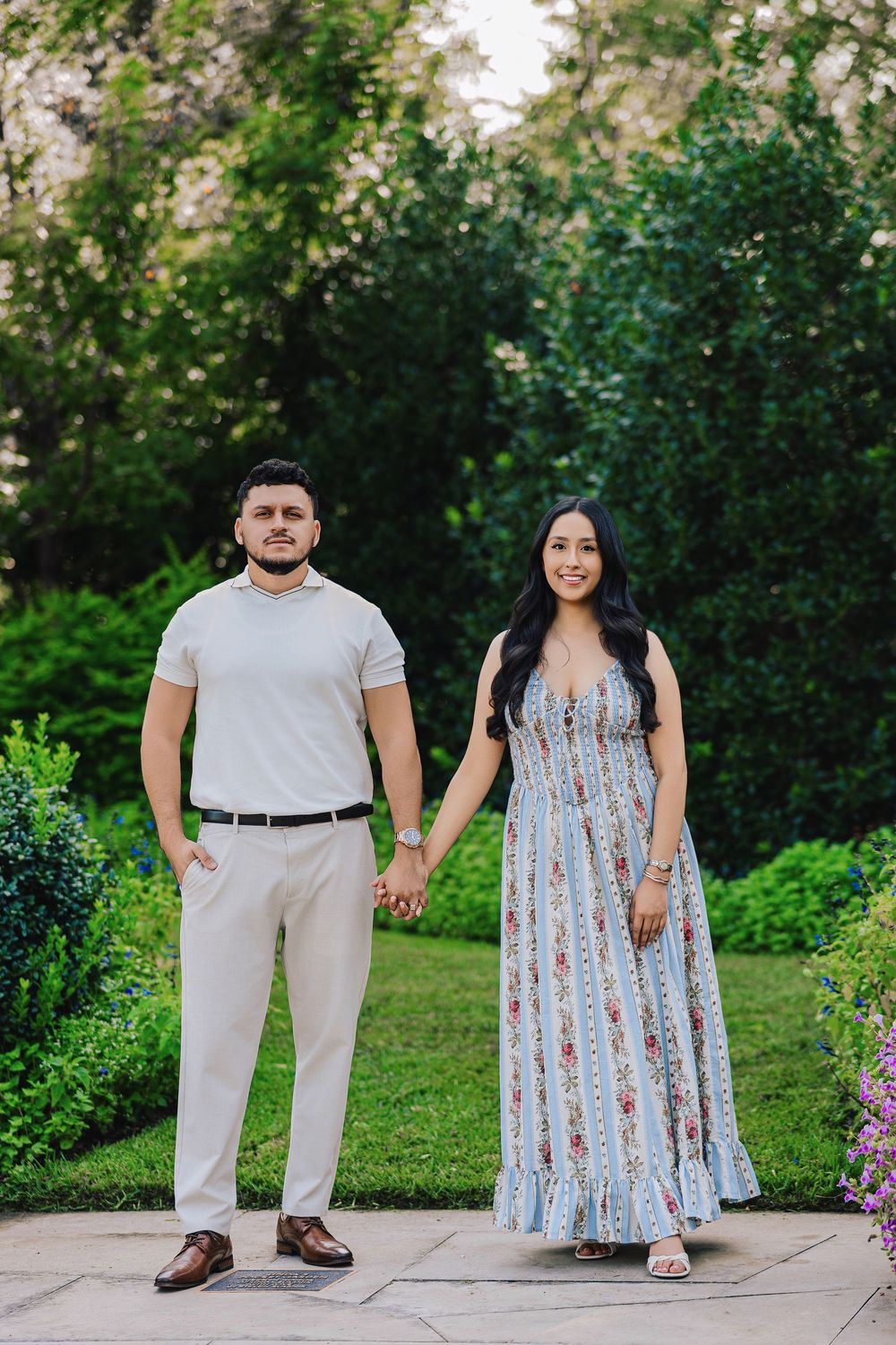 Couple holding hands in a lush garden, man in white shirt and khakis, woman in floral maxi dress.