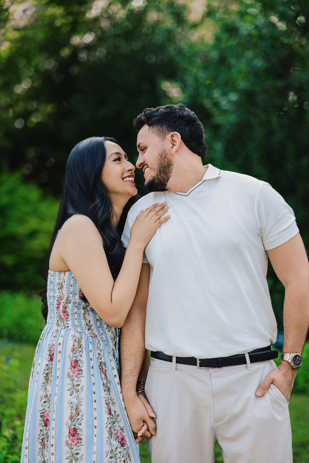 A smiling couple holds hands outdoors, the woman in a floral dress rests her hand on the man's chest in a lush green park.