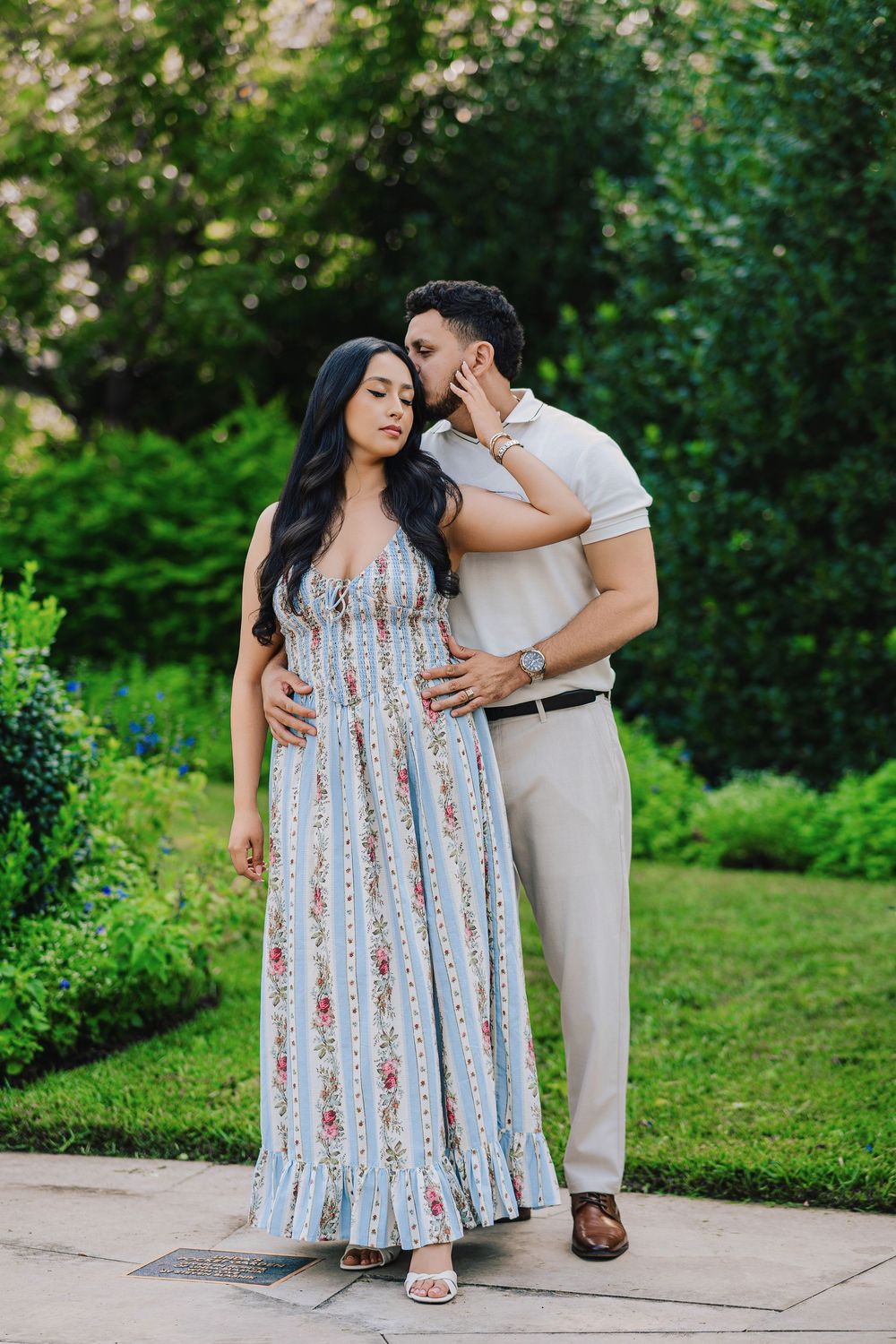 A man kisses a pregnant woman in a floral blue dress outdoors in a lush green garden setting.